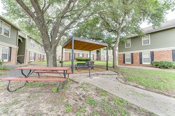A picnic table is in the middle of a grassy area with a pavilion and apartment buildings in the background.